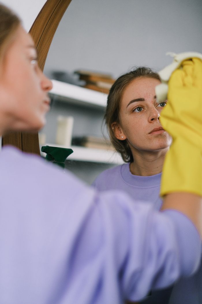 services-02 Crop female in purple sweatshirt and latex gloves washing mirror in light apartment in daytime